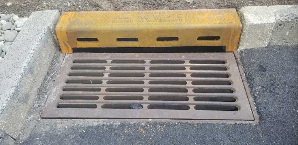 Street drain with metal grate beside a yellow curb block on a concrete roadway