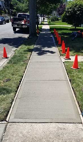 Sidewalk path bordered by orange cones beside a grassy street and parked car.