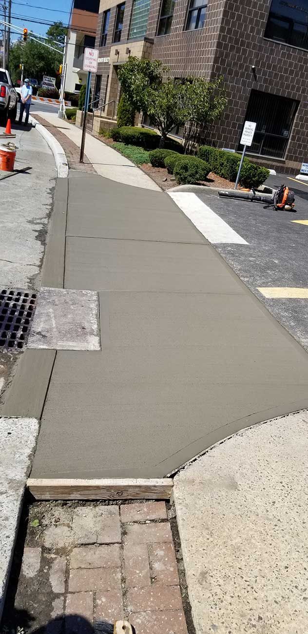 Concrete sidewalk ramp leading along a brick building with orange cones and landscaping on a sunny street