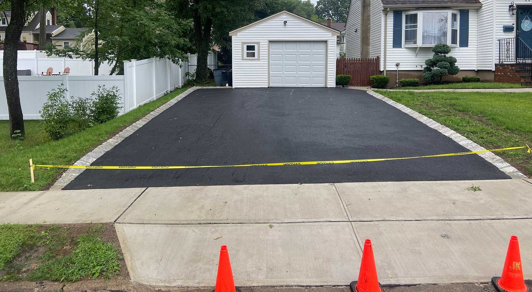 Driveway with fresh black asphalt blocked by yellow tape, orange cones in front, house and garage in background.