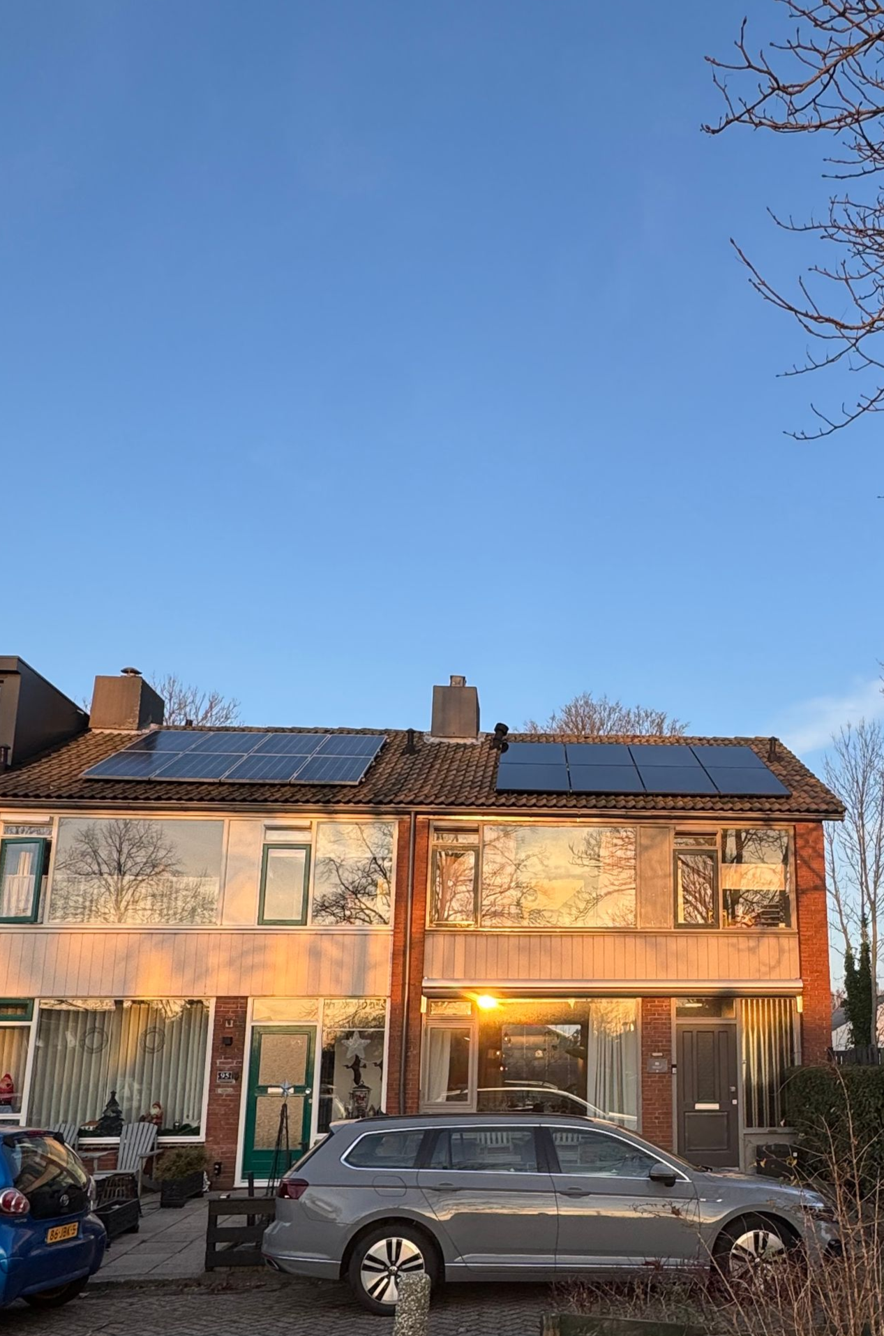 Twee verdiepingen tellende rijtjeshuizen met zonnepanelen op het dak, een auto ervoor geparkeerd, verlicht door de zonsondergang.