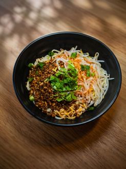 Bowl of noodles topped with minced meat, herbs, and shredded vegetables on a wooden table