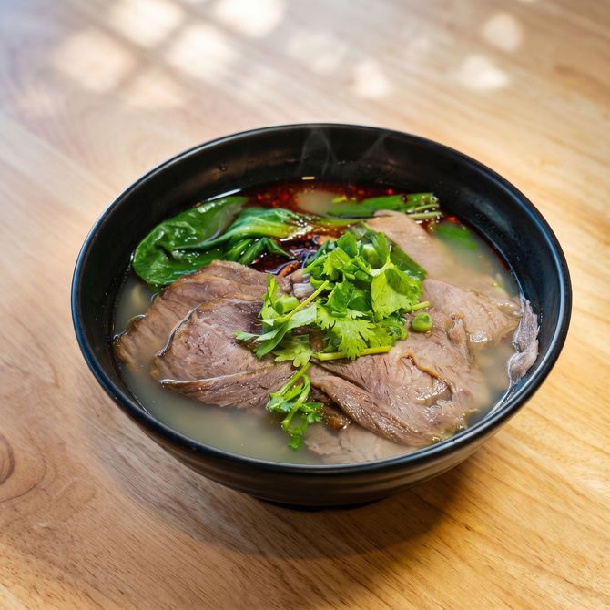 Bowl of beef noodle soup topped with greens on a wooden table