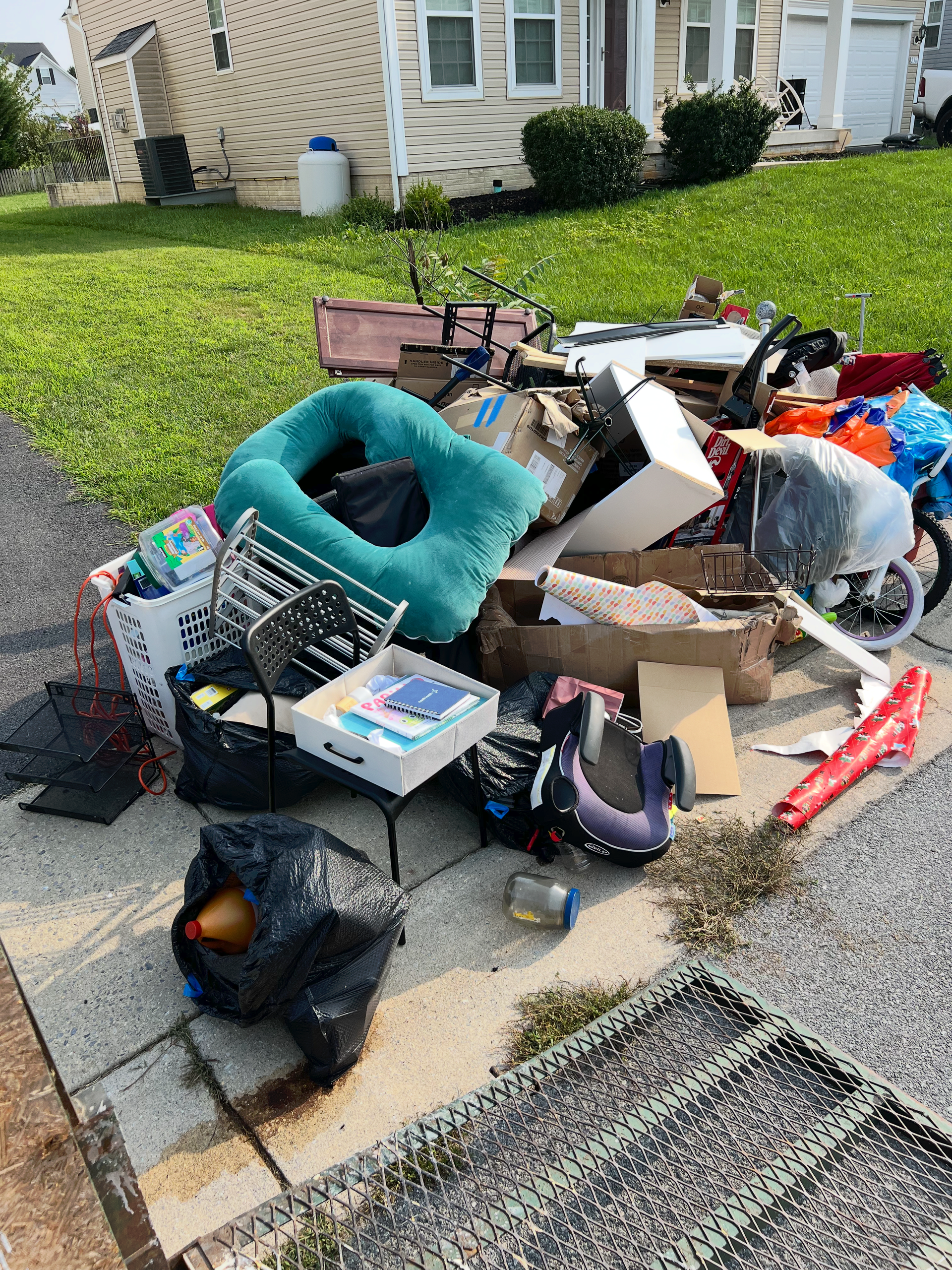 Pile of discarded furniture, boxes, and trash bags piled on a sidewalk beside a house and lawn.