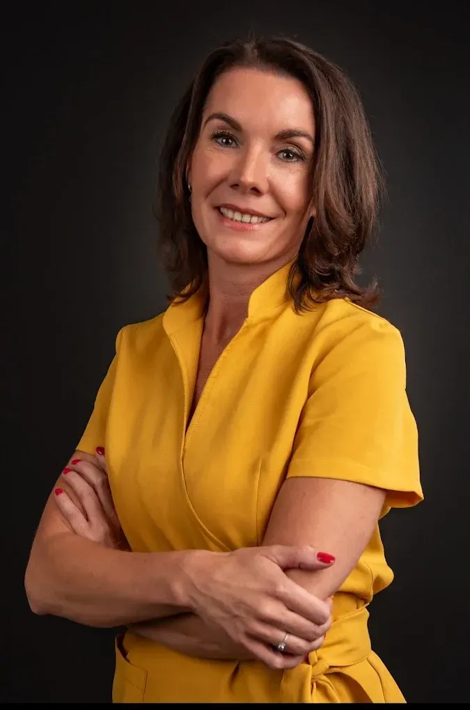 Vrouw in een gele jurk met gekruiste armen, glimlachend tegen een donkere studioachtergrond.