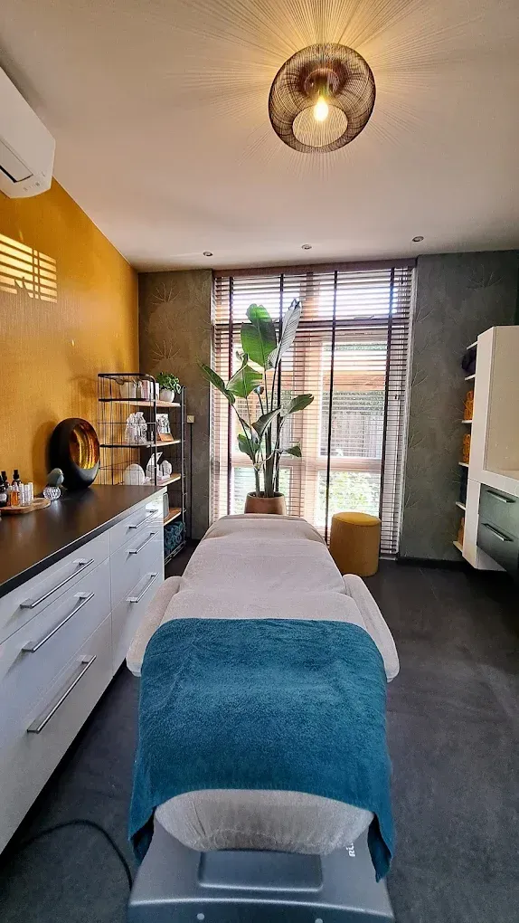 Massageruimte met behandeltafel, houten kasten, plant en veel daglicht door het raam.