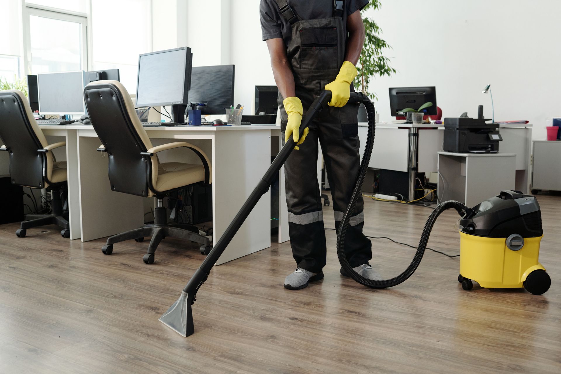 Man in work clothes vacuuming a wooden floor in an office, using a yellow and black vacuum cleaner.