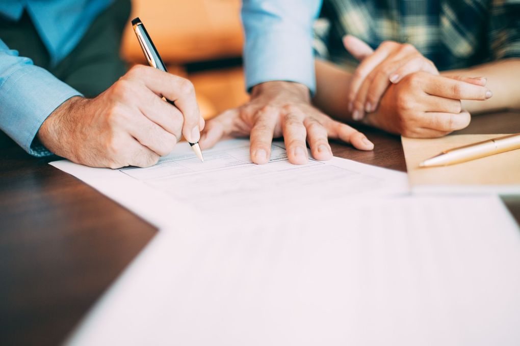 Person signing a document at a table; another person’s hand rests on their arm.