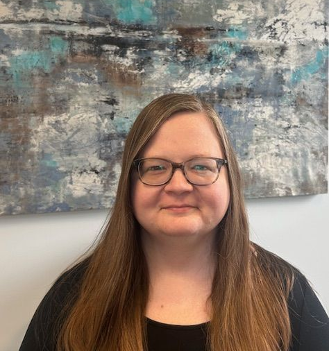 Woman with glasses and long brown hair smiles in front of abstract blue, grey, and white art.