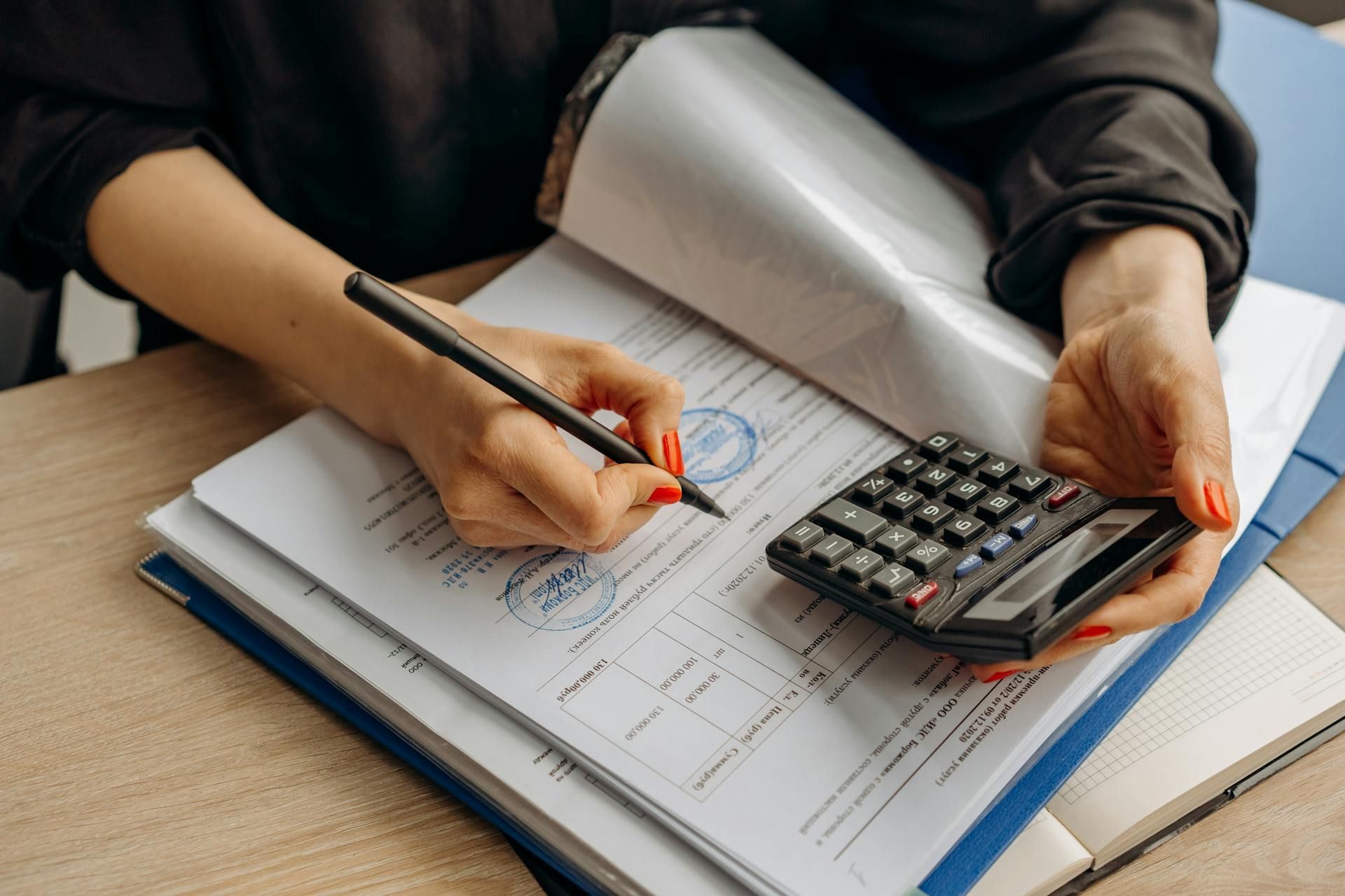 Woman's hands writing on documents with a pen, calculator on desk.