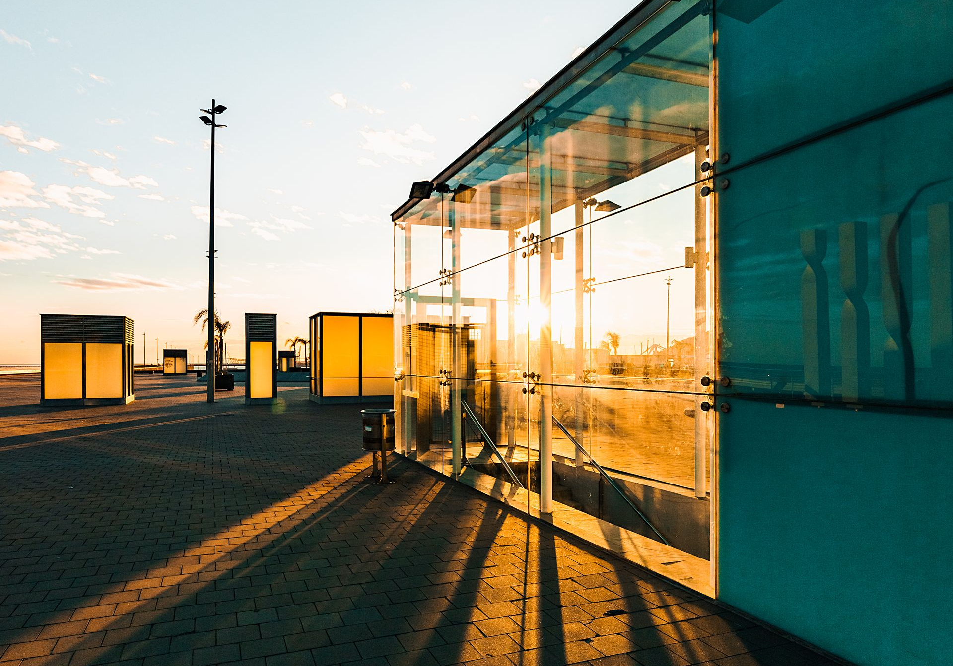 A modern, glass-walled building reflects the setting sun; other yellow structures are in the distance, shadows cast on the ground.