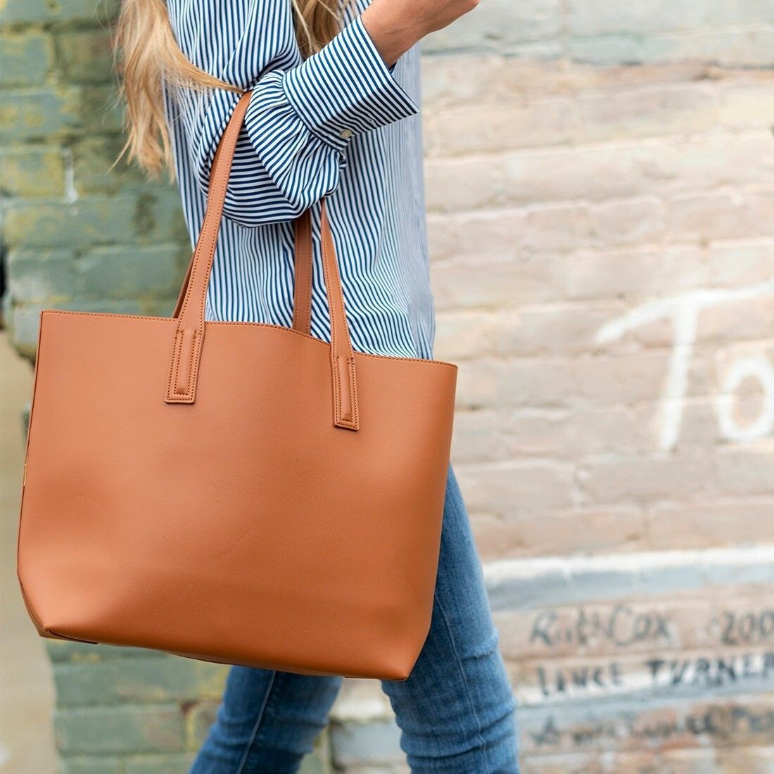 A woman is carrying a brown tote bag in front of a brick wall.