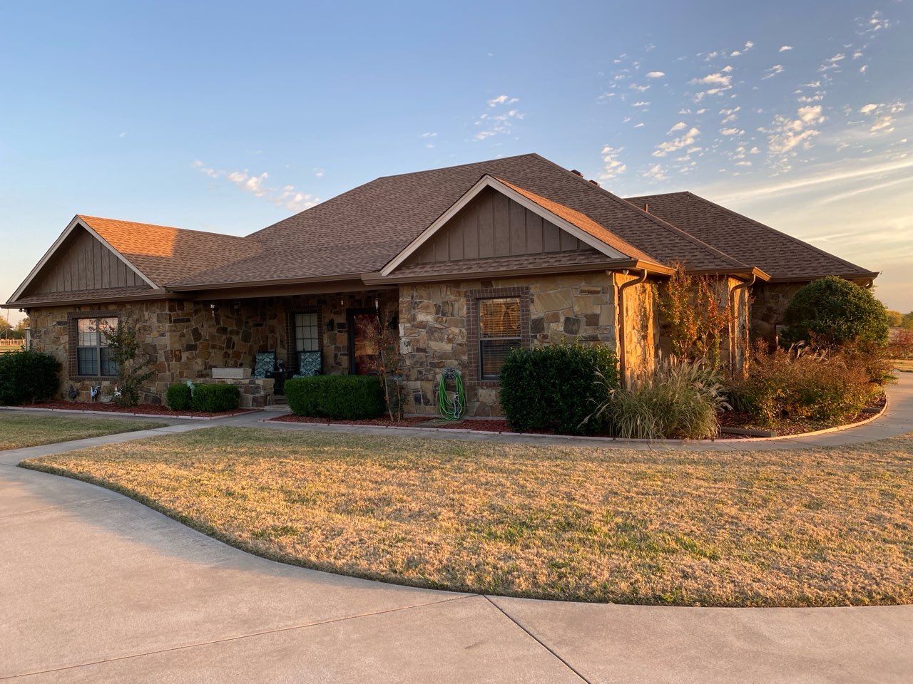 Stone-fronted ranch house with brown roof, green bushes, and a curved driveway under a blue and orange sky.