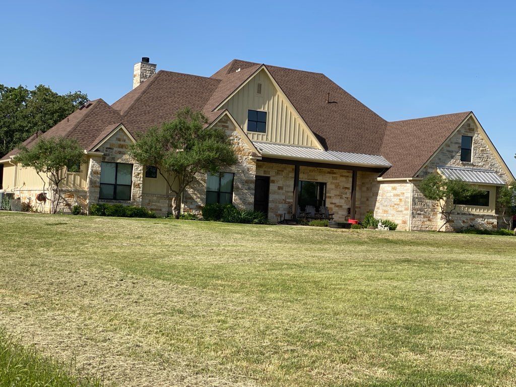 Stone and tan house with brown roof and porch on a grassy lawn under a blue sky.