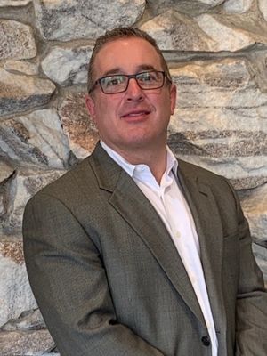 Man in glasses and gray blazer standing against a stone wall, smiling slightly.