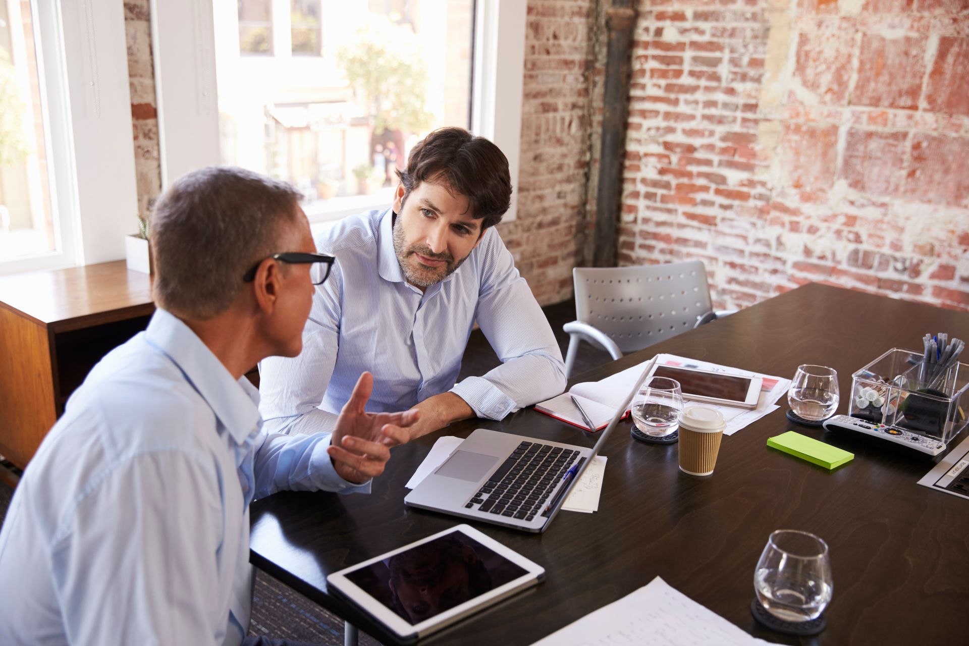 Two people discussing business at a conference table with laptops, papers, and glasses in a brick-walled office