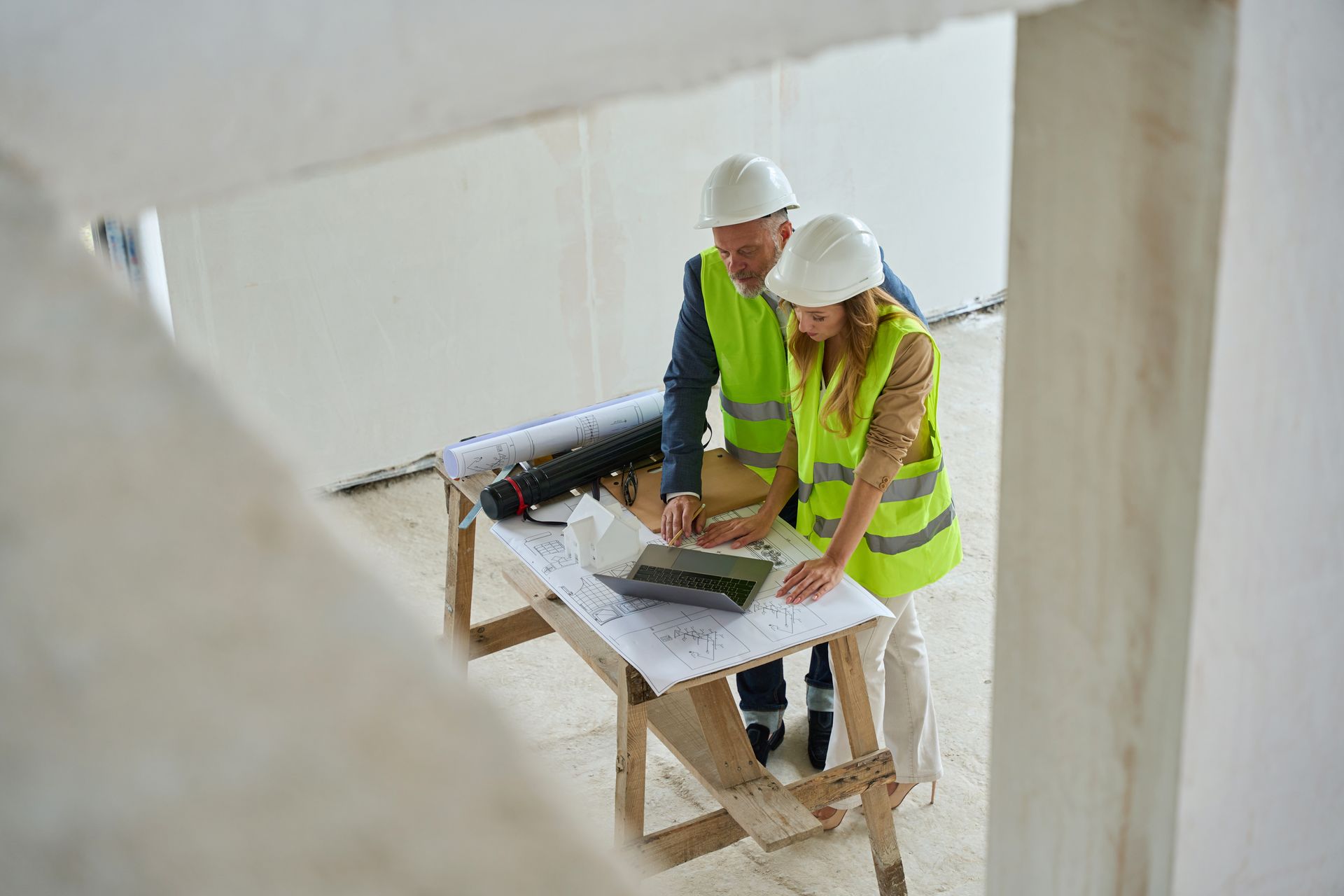 Two construction workers in hard hats and safety vests reviewing plans on a scaffold indoors