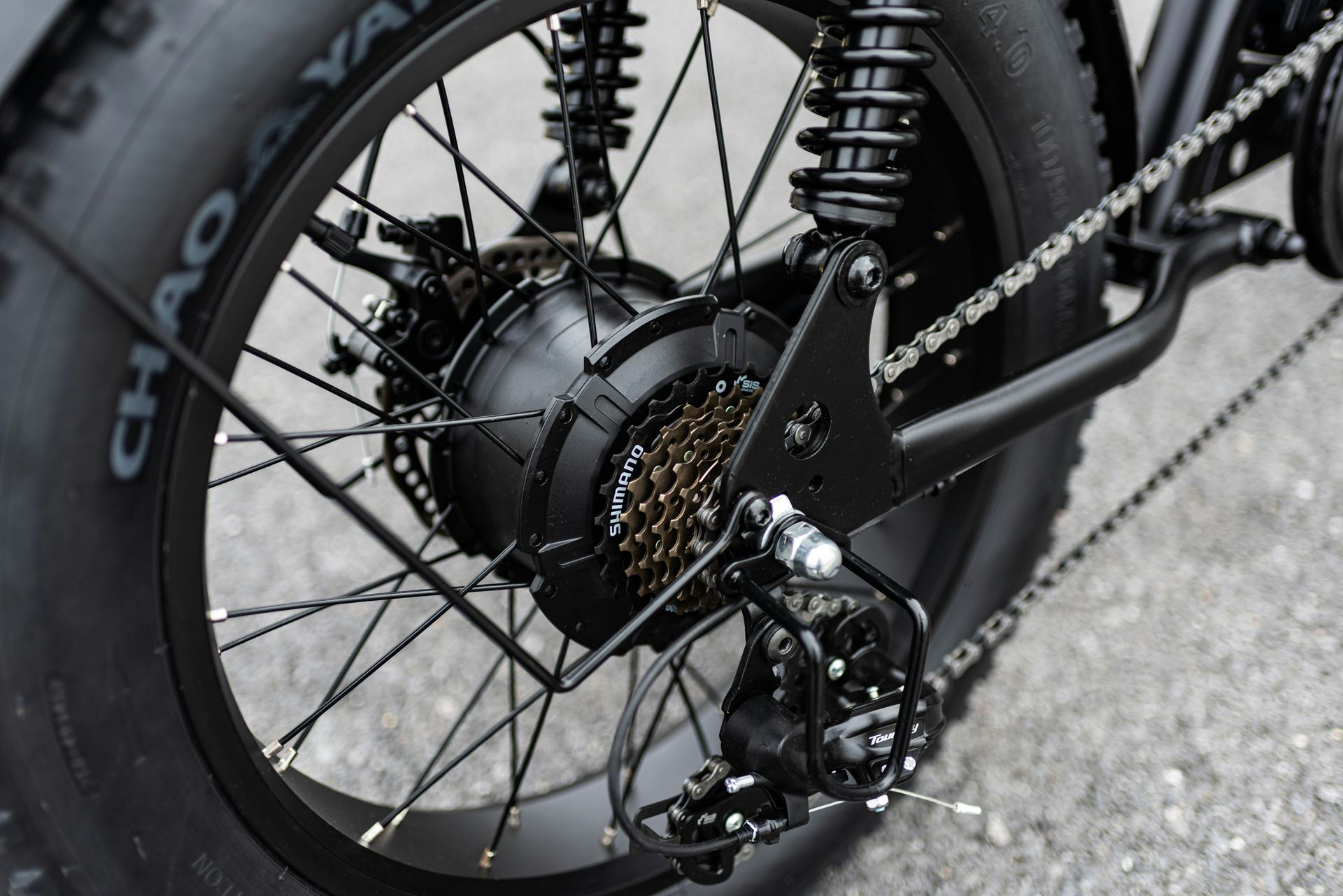 Close-up van het achterwiel, de motor, de ketting en de vering van een zwarte elektrische fiets op de stoep.