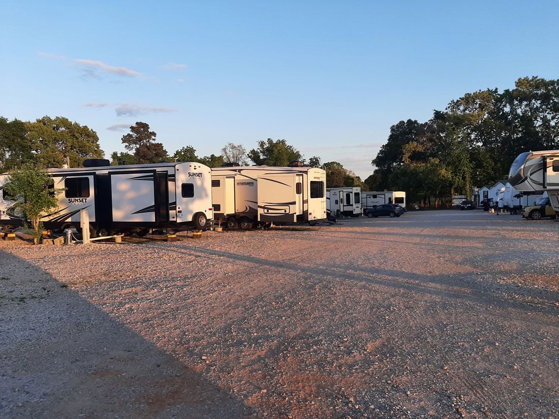 RVs parked in a gravel lot under a clear, blue sky. Trees line the edges.