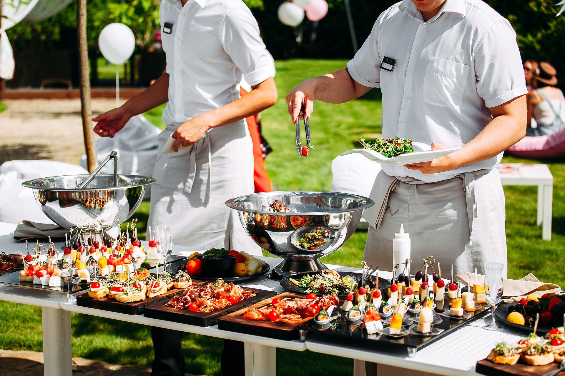 Catering buffet with chefs serving appetizers and salad outdoors on a sunny lawn.