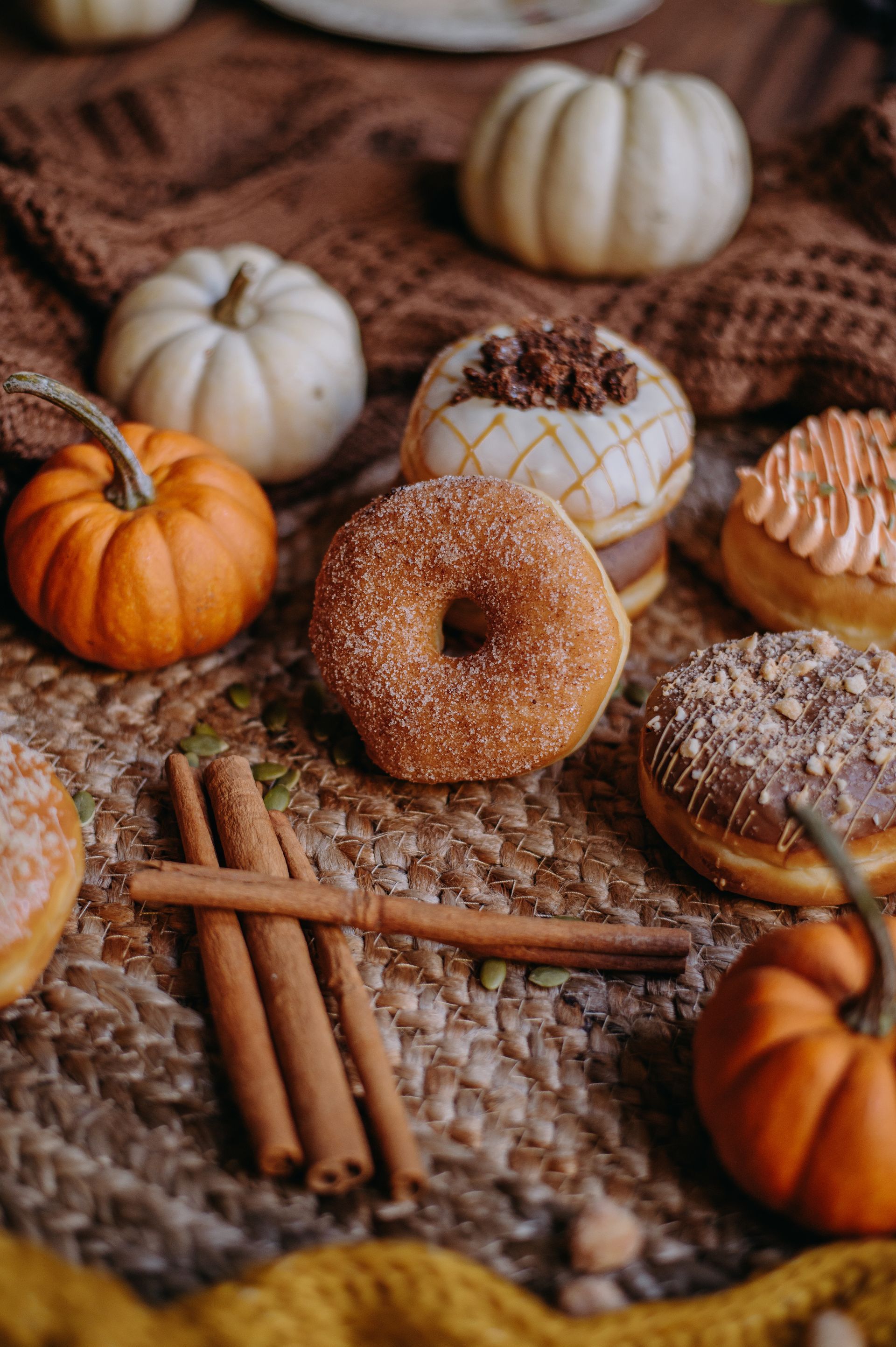 Fall donuts, cinnamon sticks, and small pumpkins on a woven mat with cozy brown fabric backdrop