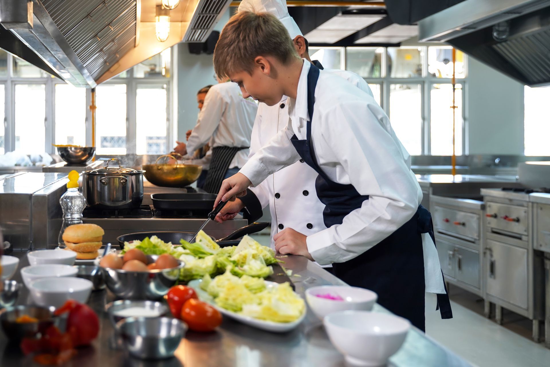 Chef preparing vegetables in a stainless-steel kitchen with cooking pots and fresh produce nearby