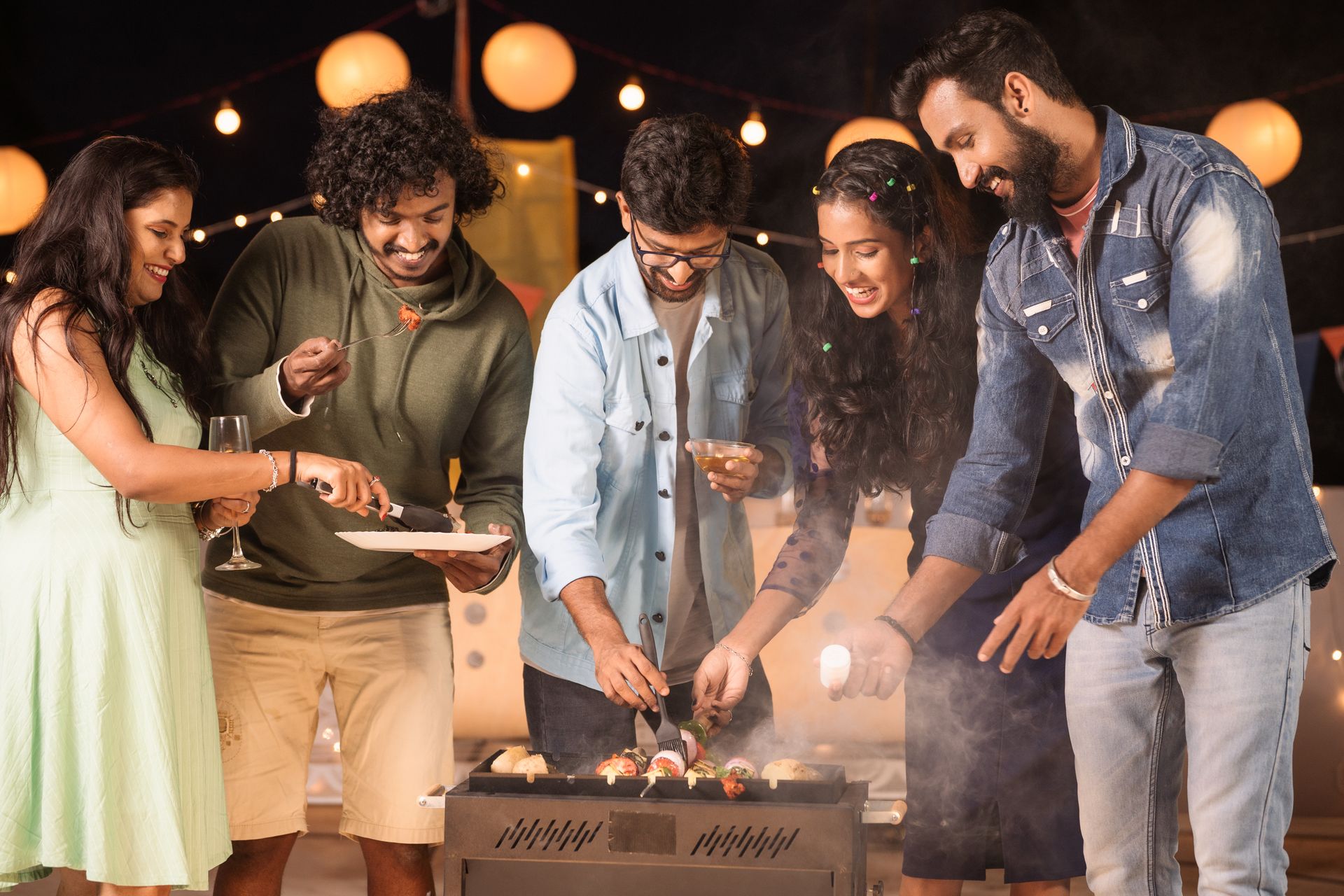 Friends gathered around a lit tabletop grill at an outdoor evening party, smiling and cooking food together.