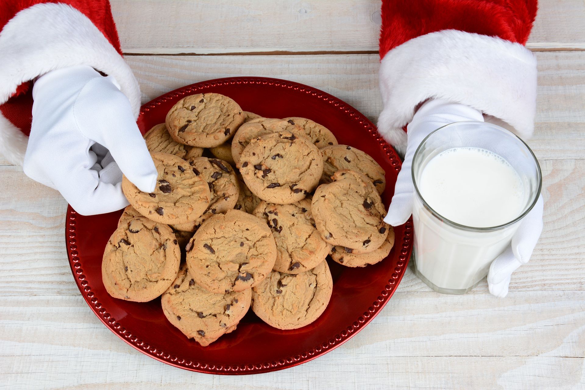 Santa-gloved hand holds cookie over red plate with cookies and a glass of milk