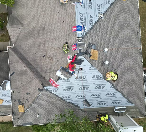 Roofers working on a roof. Brown shingles, blue underlayment, safety gear, and tools are visible.