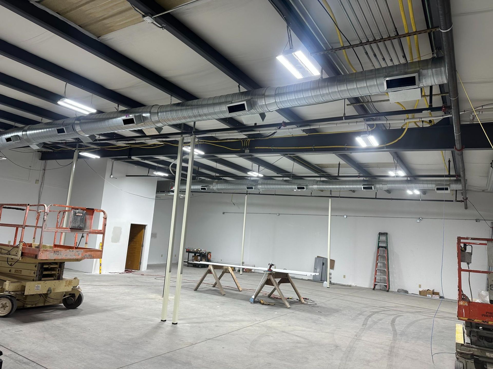 Partially finished industrial interior with exposed ceiling, ductwork, ladders, and scissor lifts on a concrete floor