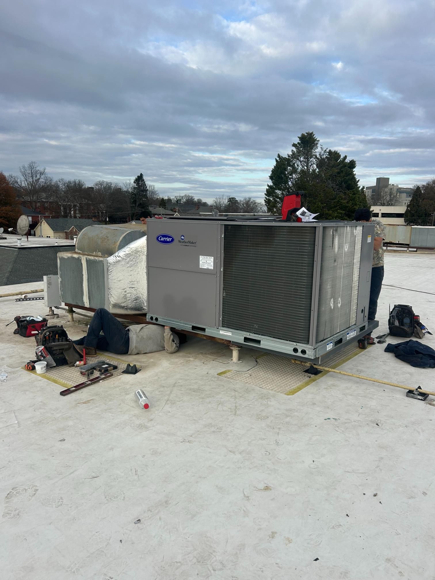 HVAC unit on a rooftop with two workers and scattered tools under a cloudy sky
