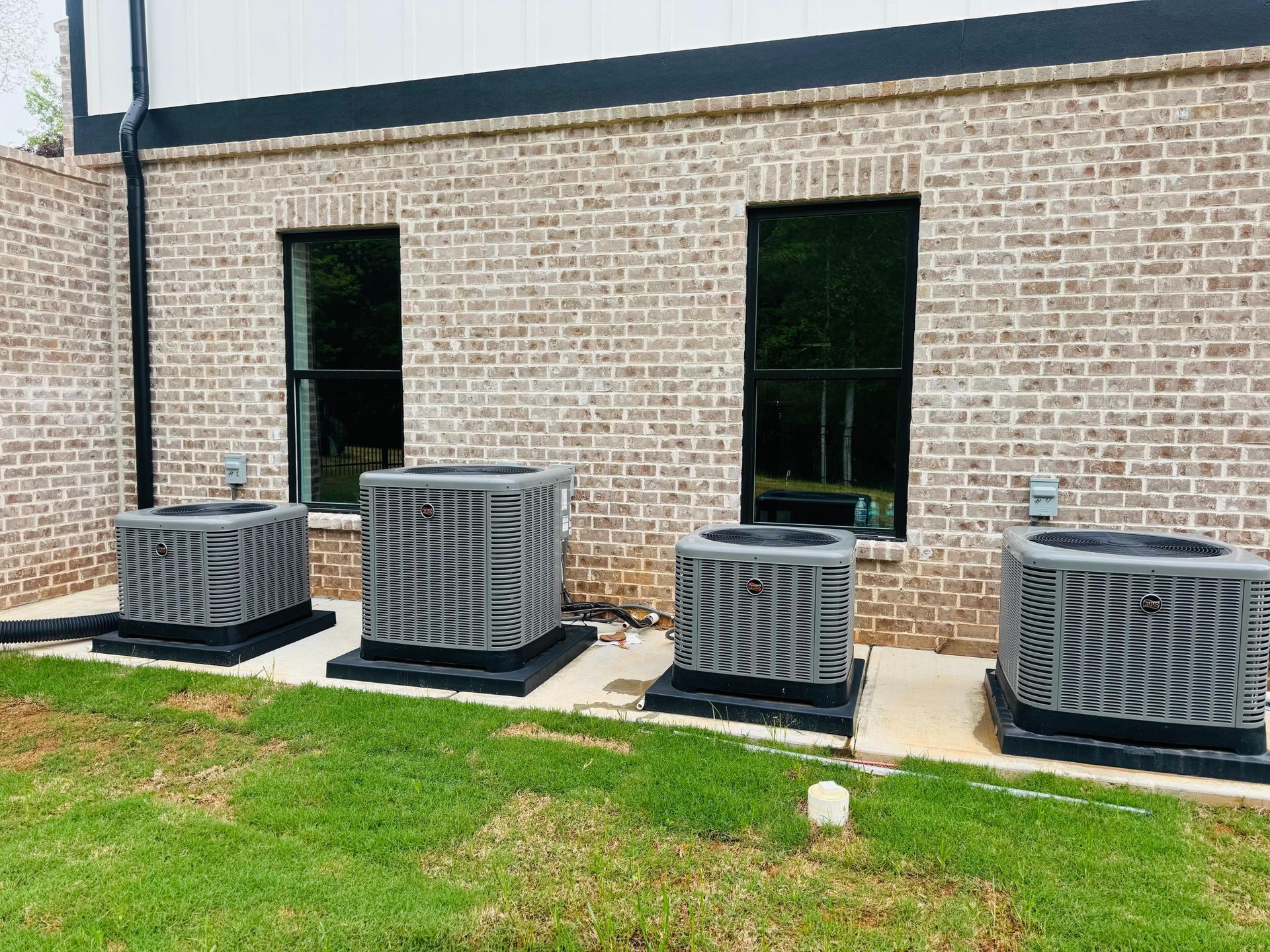 Four gray outdoor HVAC units beside a brick building with two windows and grass in front.