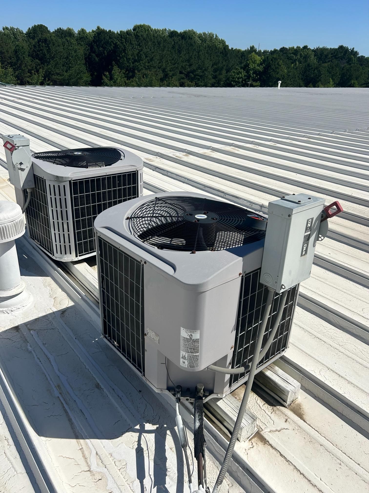 Two rooftop HVAC units on a white metal roof with trees in the background