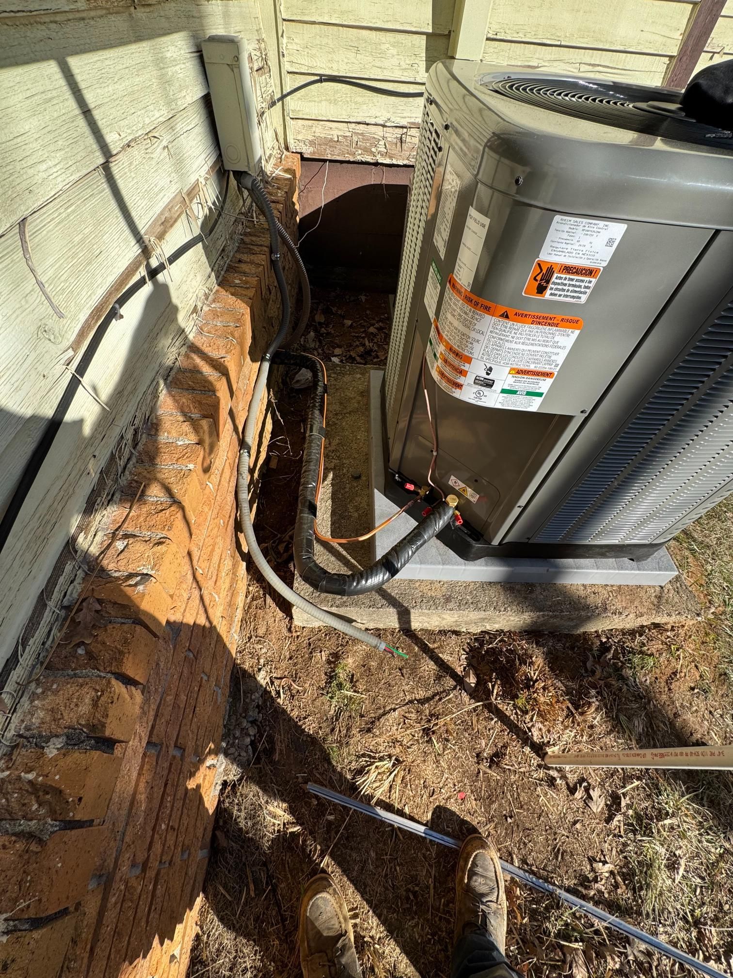 Outdoor HVAC unit beside a house wall with exposed pipes and dirt trenching around the foundation
