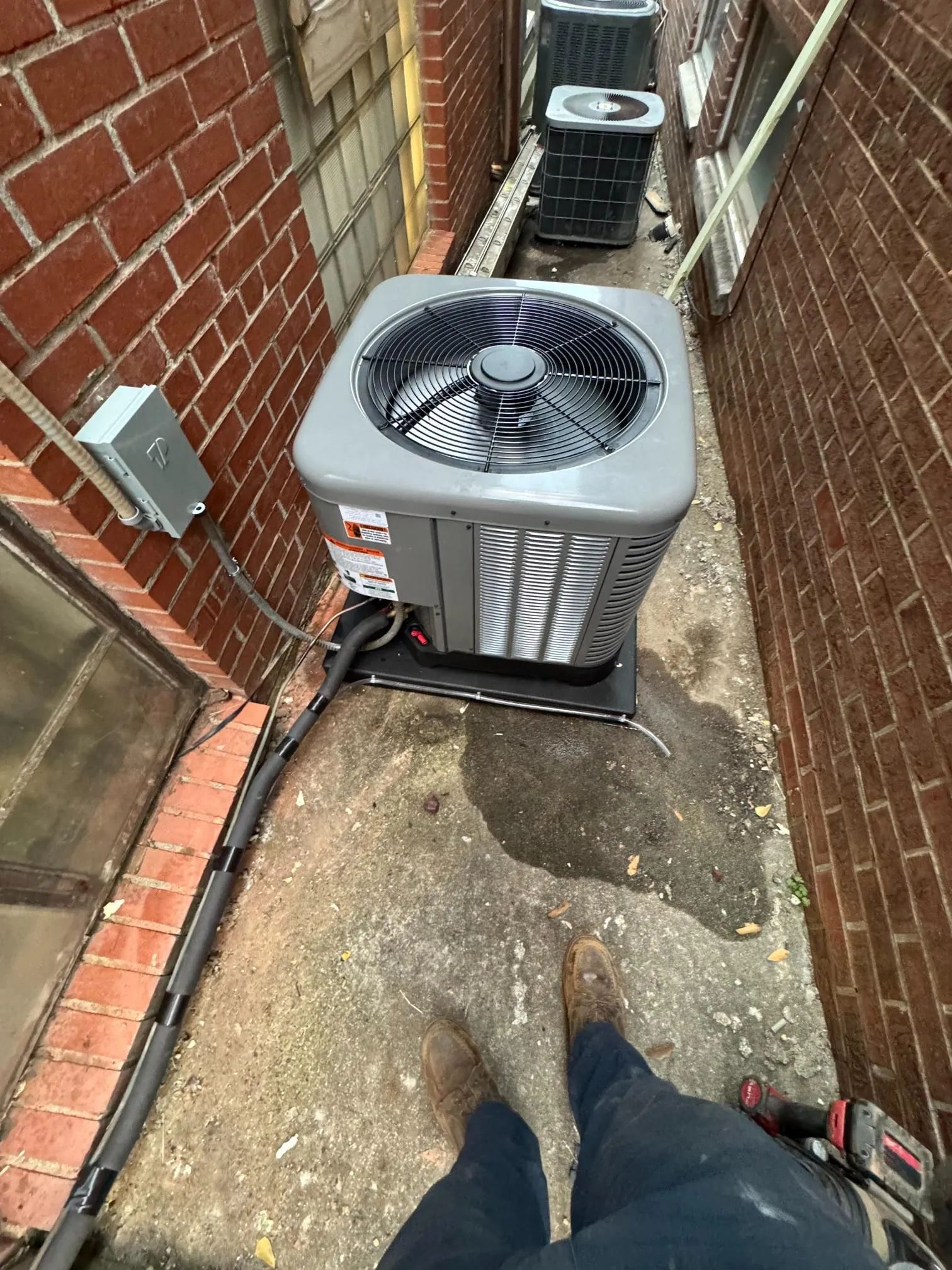 Outdoor air conditioner unit in a narrow alley beside a brick wall, with a person’s legs visible in the foreground.