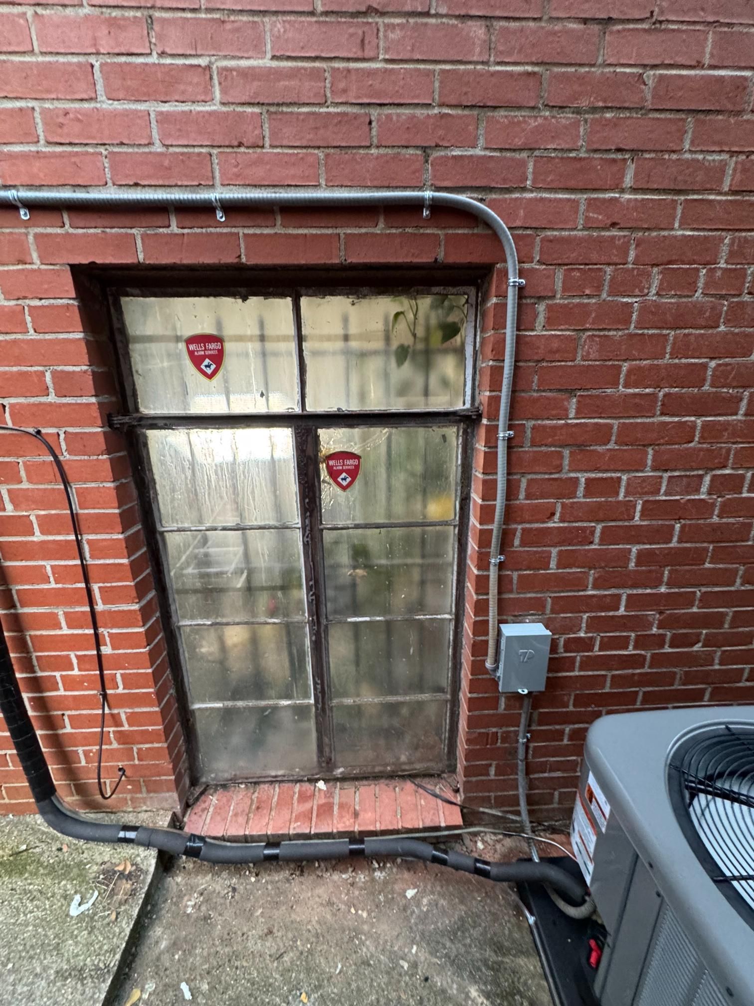 Small basement window in a red brick wall with metal bars and an exterior AC unit nearby.