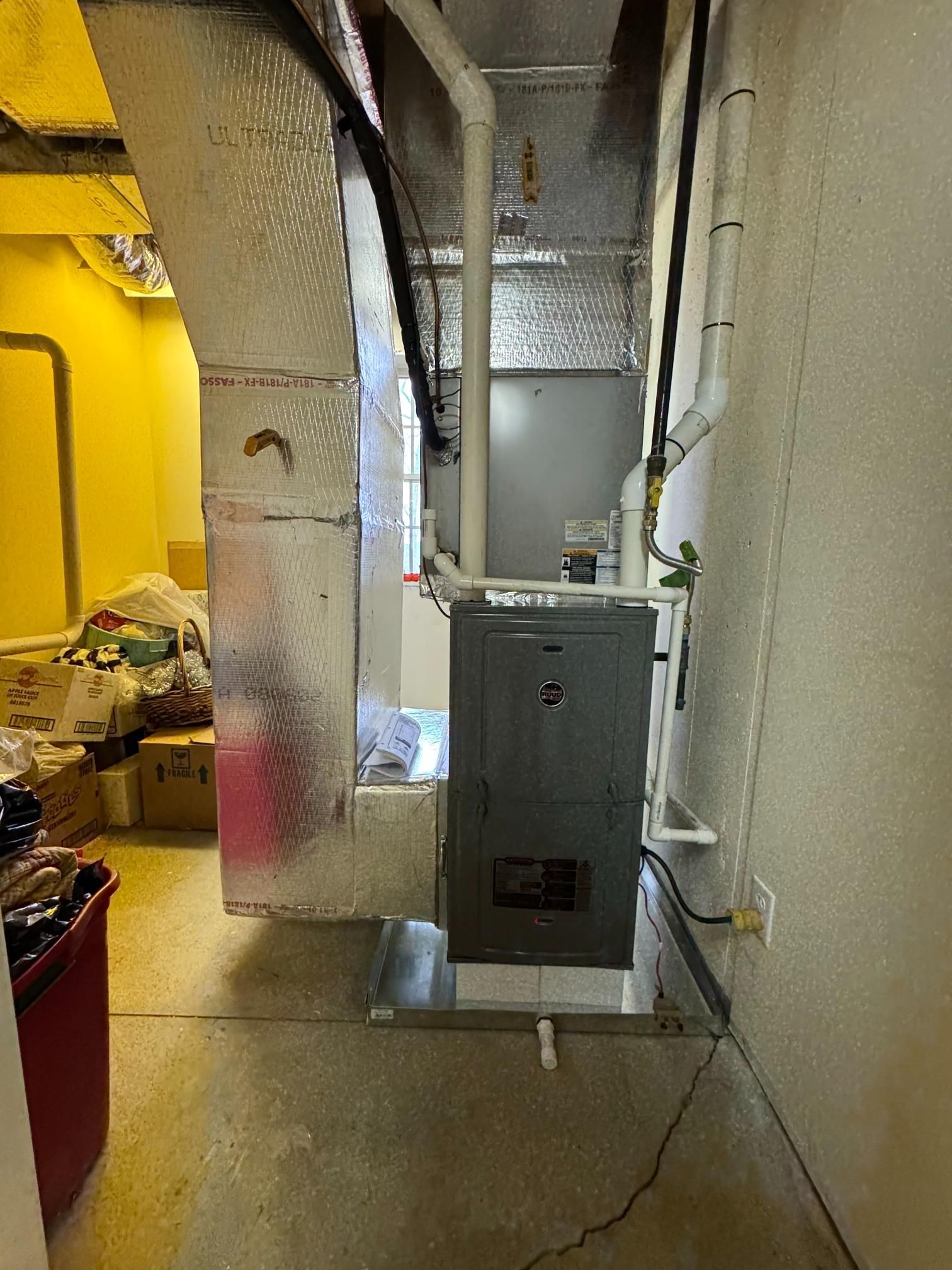 Basement utility room with gray furnace, silver ducts, yellow wall, and exposed pipes.