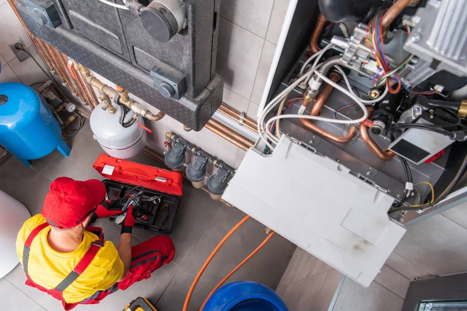 Plumber in red coveralls working on a furnace with tools, inside a utility room.