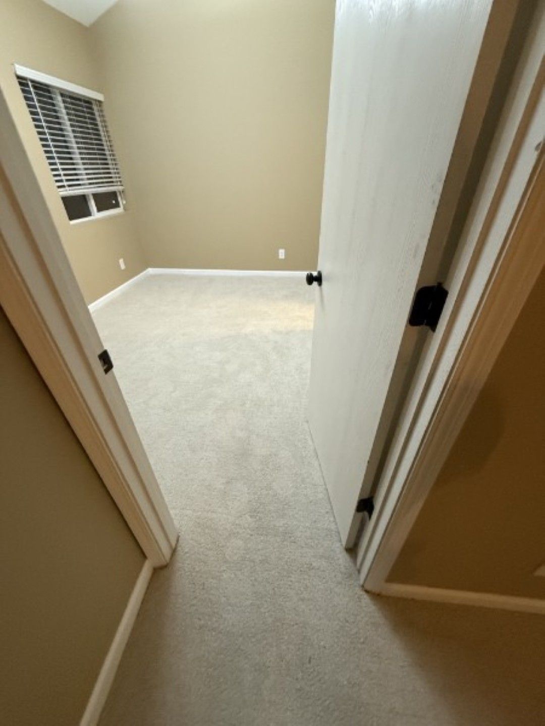 Open doorway to an empty beige bedroom with carpet and a window blinds.