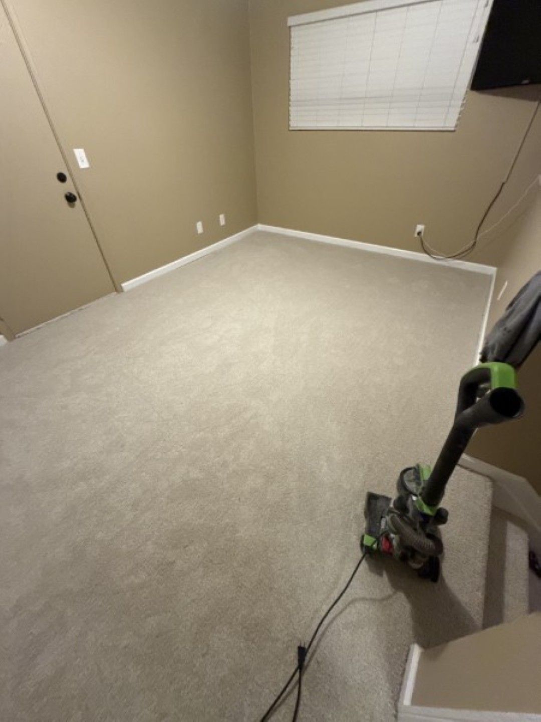 Empty beige bedroom with carpet, window blinds, and a vacuum cleaner on the right side.