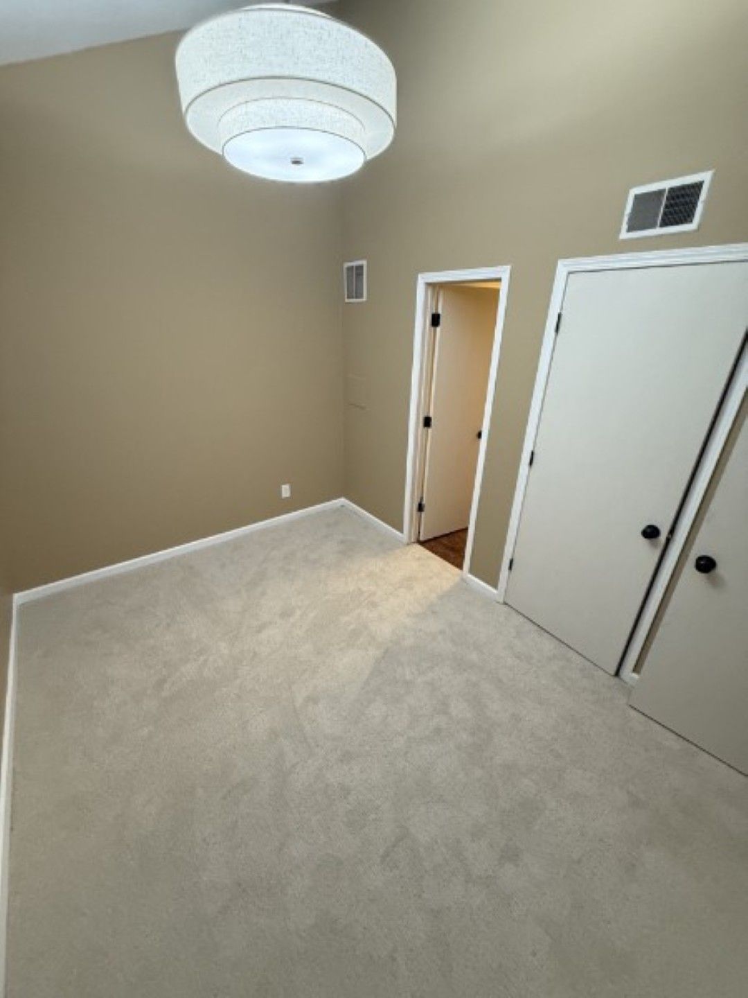 Empty beige bedroom with carpet, white closet doors, and a ceiling light fixture.