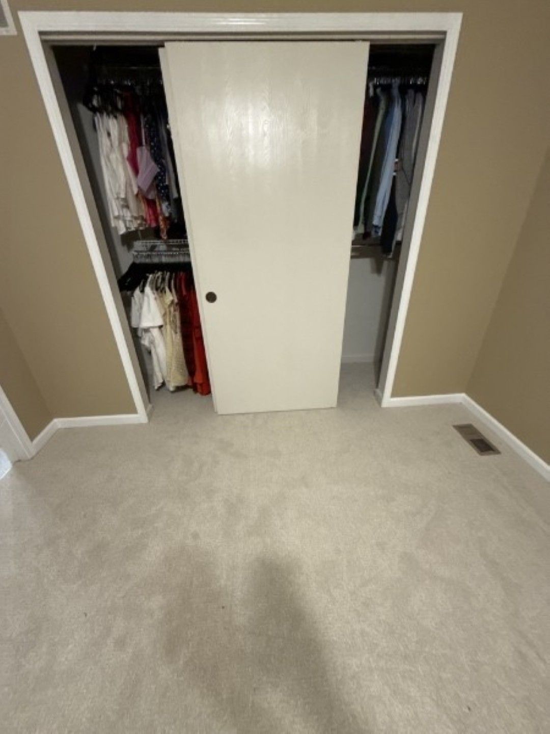 Open closet with sliding white doors, hanging clothes, and beige carpet in a bedroom.