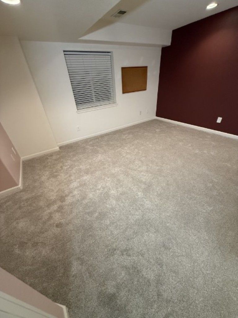 Empty carpeted room with sloped ceiling, white walls, maroon accent wall, window, and corkboard.