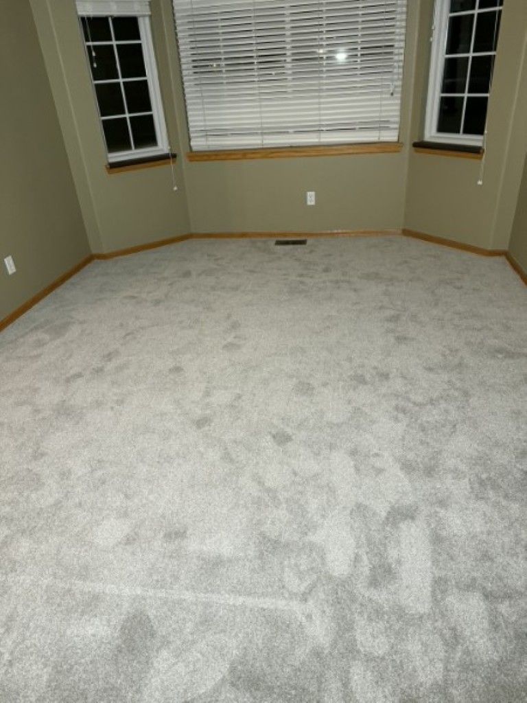 Empty beige carpeted room with bay window and blinds, taupe walls, and wood trim