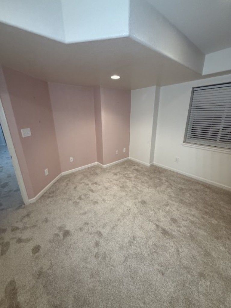 Empty beige carpeted room with pink accent walls, recessed light, and a window with blinds