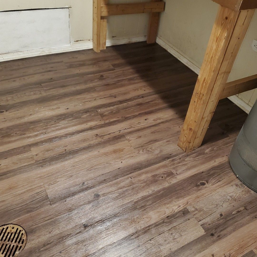 Wood-look floor with a round drain, table legs, and a gray container in a corner of a room