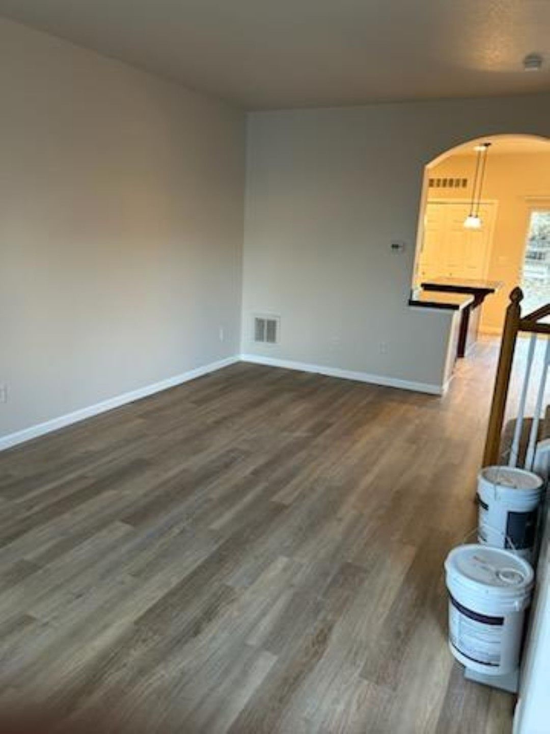 Empty room with wood-look flooring, white walls, and an arched doorway to a lit kitchen area.