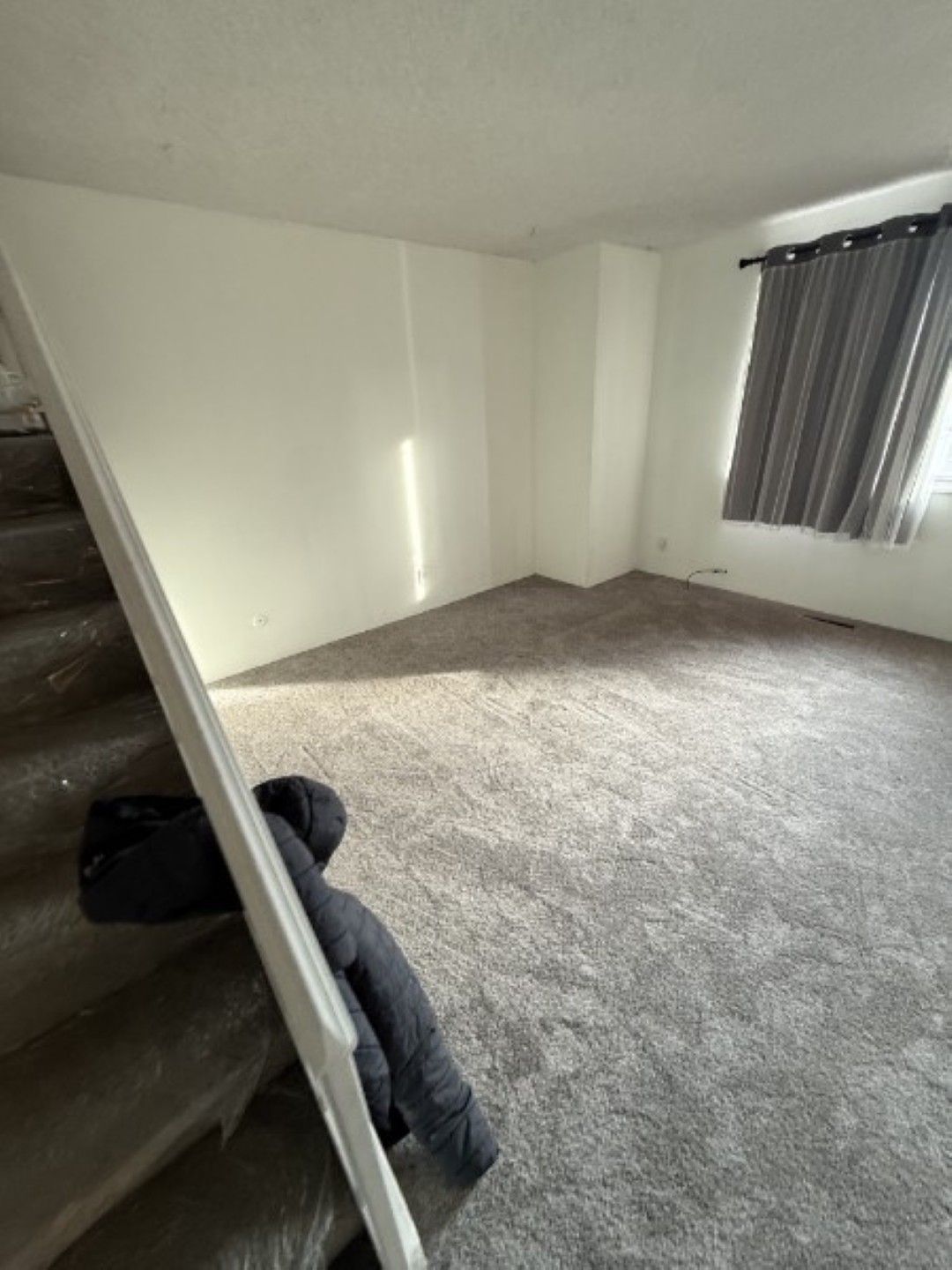 Empty carpeted room with white walls, staircase on the left, and a window with dark curtains.
