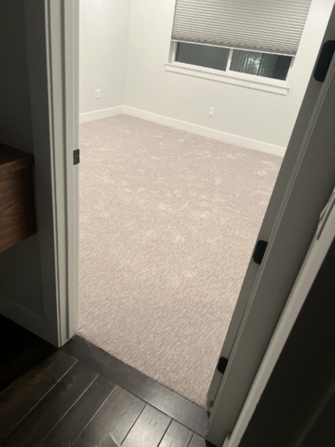Open doorway to an empty beige-carpeted room with a window and dark wood floor outside the door