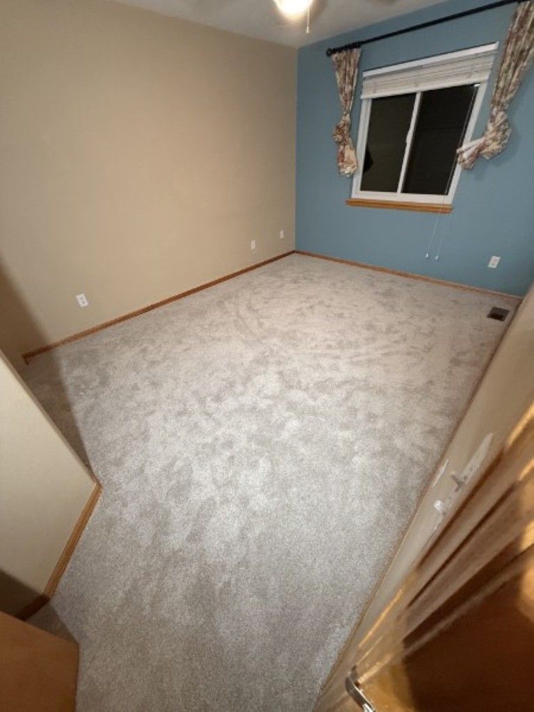 Empty carpeted bedroom with beige walls, blue accent wall, and a window with curtains.